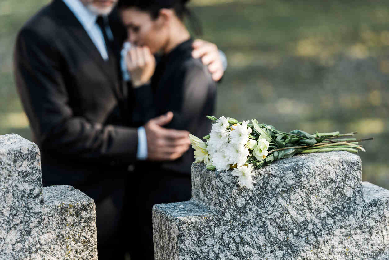 A man in a suit comforts a woman in a black shirt, who is crying at a grave with a bouquet of white flowers on the headstone for someone's wrongful death